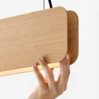 nina-japandi-linear-oak-suspension-lamp Close-up detail of the Nina linear pendant light by Singular Lighting, featuring natural oak wood panels, black suspension cables, and a warm LED glow, gently held by a hand.