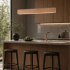Singular Lighting - NINA Pendant P120-04B A modern kitchen island with a natural oak pendant light (the 'Nina' lamp) hanging above it. The island has dark wooden stools, a bowl of pears, and a vase, with a leafy window view.
