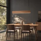 Singular Lighting - NINA Pendant P120-06B A sleek natural oak pendant light (the 'Nina' lamp) hanging above a modern wooden dining table and grey-upholstered chairs. Sunlight from a large window creates intricate shadow patterns on the wall