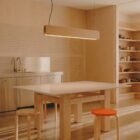 Singular Lighting - NINA Pendant P120-16B A minimalist, plywood kitchen and workspace illuminated by a natural oak pendant light (the 'Nina' lamp). A wooden table, two simple plywood stools, one with an orange seat, and exposed shelves are in the space.