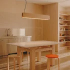 Singular Lighting - NINA Pendant P120-16B A minimalist, plywood kitchen and workspace illuminated by a natural oak pendant light (the 'Nina' lamp). A wooden table, two simple plywood stools, one with an orange seat, and exposed shelves are in the space.