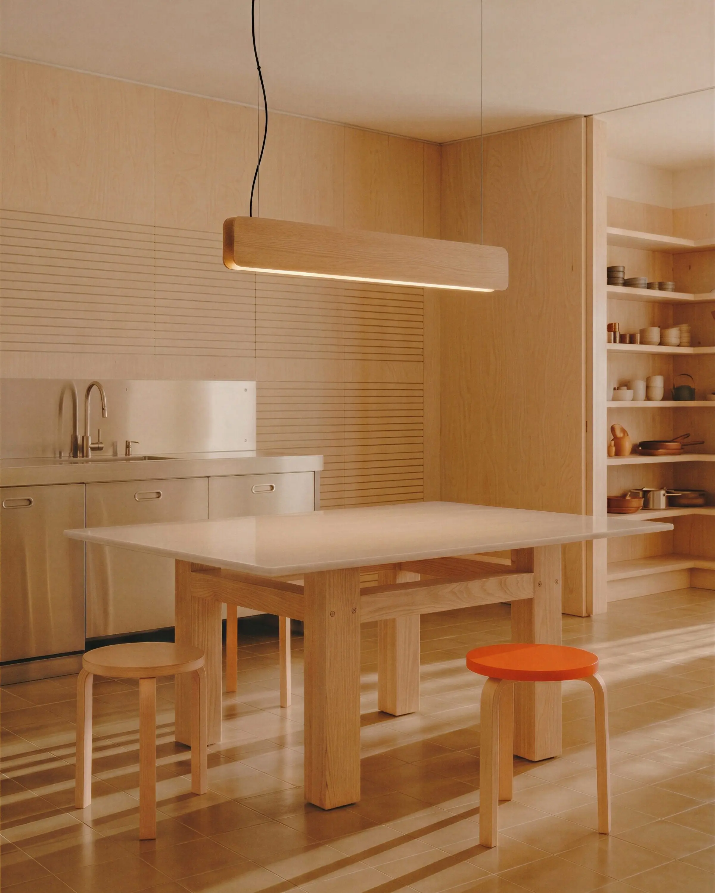Singular Lighting - NINA Pendant P120-16B A minimalist, plywood kitchen and workspace illuminated by a natural oak pendant light (the 'Nina' lamp). A wooden table, two simple plywood stools, one with an orange seat, and exposed shelves are in the space.