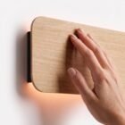 singular-lighting-nina-wall-natural-oak-wood-texture-close-up-detail Close-up detail of a hand gently touching the smooth natural oak wood finish of the Nina Wall sconce by Singular Lighting, highlighting the fine wood grain and the warm indirect light glowing from behind the rounded panel against a white wall.