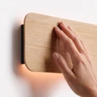 Close-up detail of a hand gently touching the smooth natural oak wood finish of the Nina Wall sconce by Singular Lighting, highlighting the fine wood grain and the warm indirect light glowing from behind the rounded panel against a white wall.