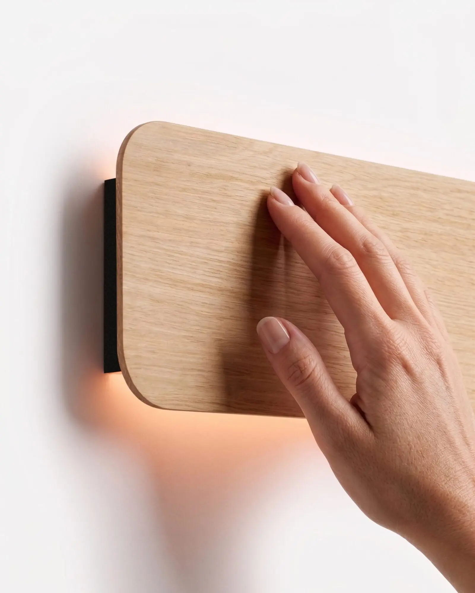Close-up detail of a hand gently touching the smooth natural oak wood finish of the Nina Wall sconce by Singular Lighting, highlighting the fine wood grain and the warm indirect light glowing from behind the rounded panel against a white wall.