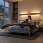 Cozy bedroom with two natural oak Nina Wall up-and-down lights, where a woman reads in bed under warm lighting. Large bed with dark bedding and concrete texture walls.