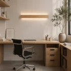 Home office with a long natural oak Nina Wall up-and-down lamp above a matching wooden desk. Ergonomic black chair, built-in bookshelves, and large window view.