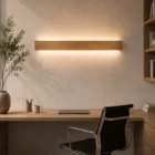 Side view of a home office desk, featuring a natural oak Nina Wall linear lamp illuminating up and down. Minimalist desk setup with laptop, plant, and office supplies.