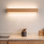 nina-wall-oak-linear-up-down-lighting-kitchen Natural oak Nina Wall lamp by Singular Lighting over a marble kitchen counter, illuminating upwards and downwards. Modern kitchen decor with wooden cabinets and utensil holder.
