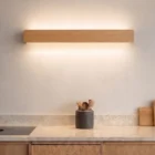 Natural oak Nina Wall lamp by Singular Lighting over a marble kitchen counter, illuminating upwards and downwards. Modern kitchen decor with wooden cabinets and utensil holder.