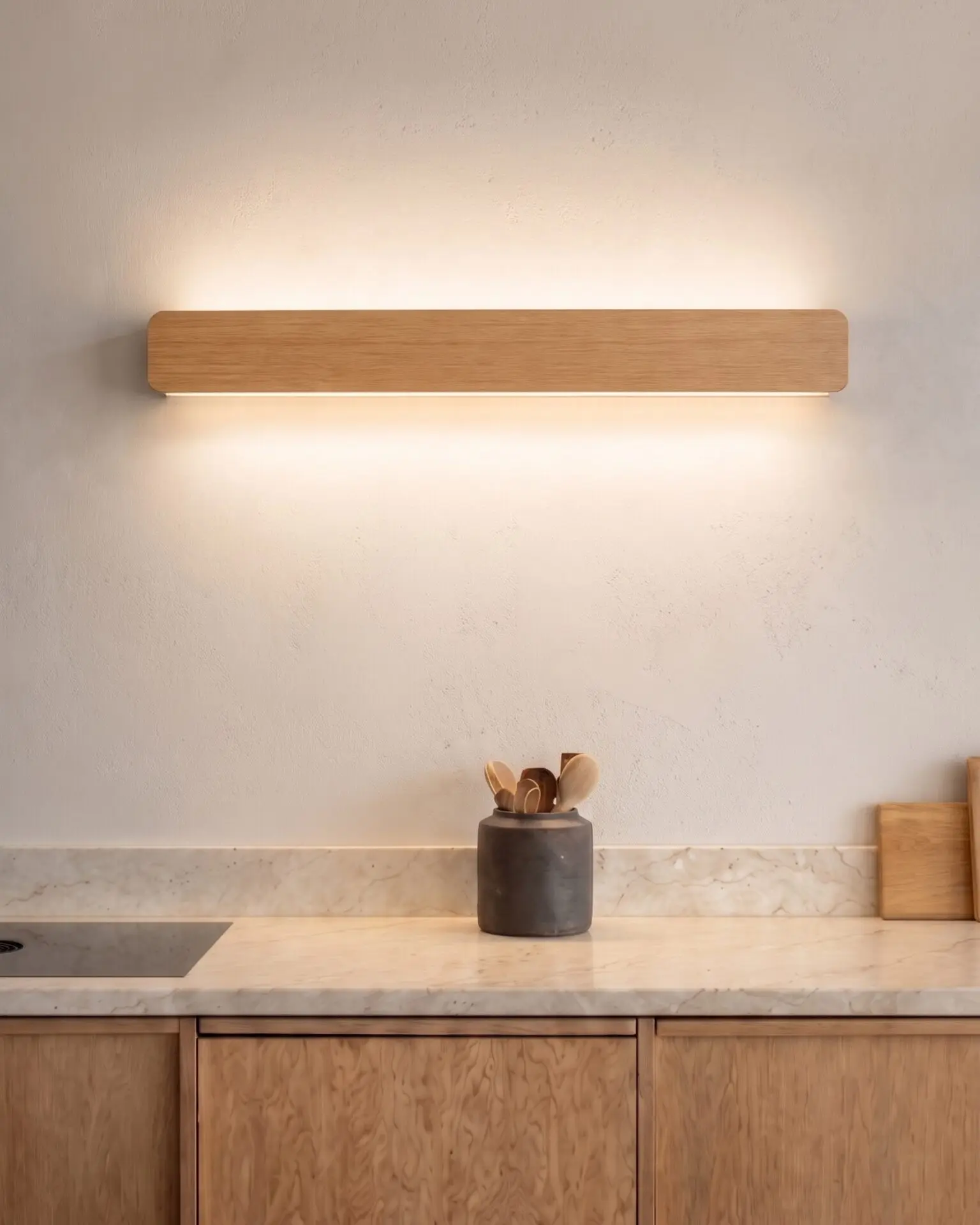 Natural oak Nina Wall lamp by Singular Lighting over a marble kitchen counter, illuminating upwards and downwards. Modern kitchen decor with wooden cabinets and utensil holder.
