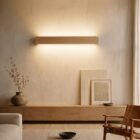 singular-lighting-nina-wall-oak-up-down-linear-sconce-living-room Singular Lighting NINA Wall W120-10B_2 natural oak linear up and down light fixture in a modern minimalist living room setting, illuminating the textured wall above a low wooden credenza, dried branches in a vase, framed art, and a leather lounge chair.