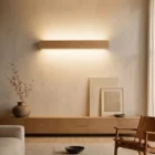 Singular Lighting NINA Wall W120-10B_2 natural oak linear up and down light fixture in a modern minimalist living room setting, illuminating the textured wall above a low wooden credenza, dried branches in a vase, framed art, and a leather lounge chair.