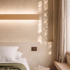 Bedside Nina Wall linear oak up-and-down light by Singular Lighting, next to a dimmer switch on a textured wall. Minimalist bedroom nightstand with a glass of water.