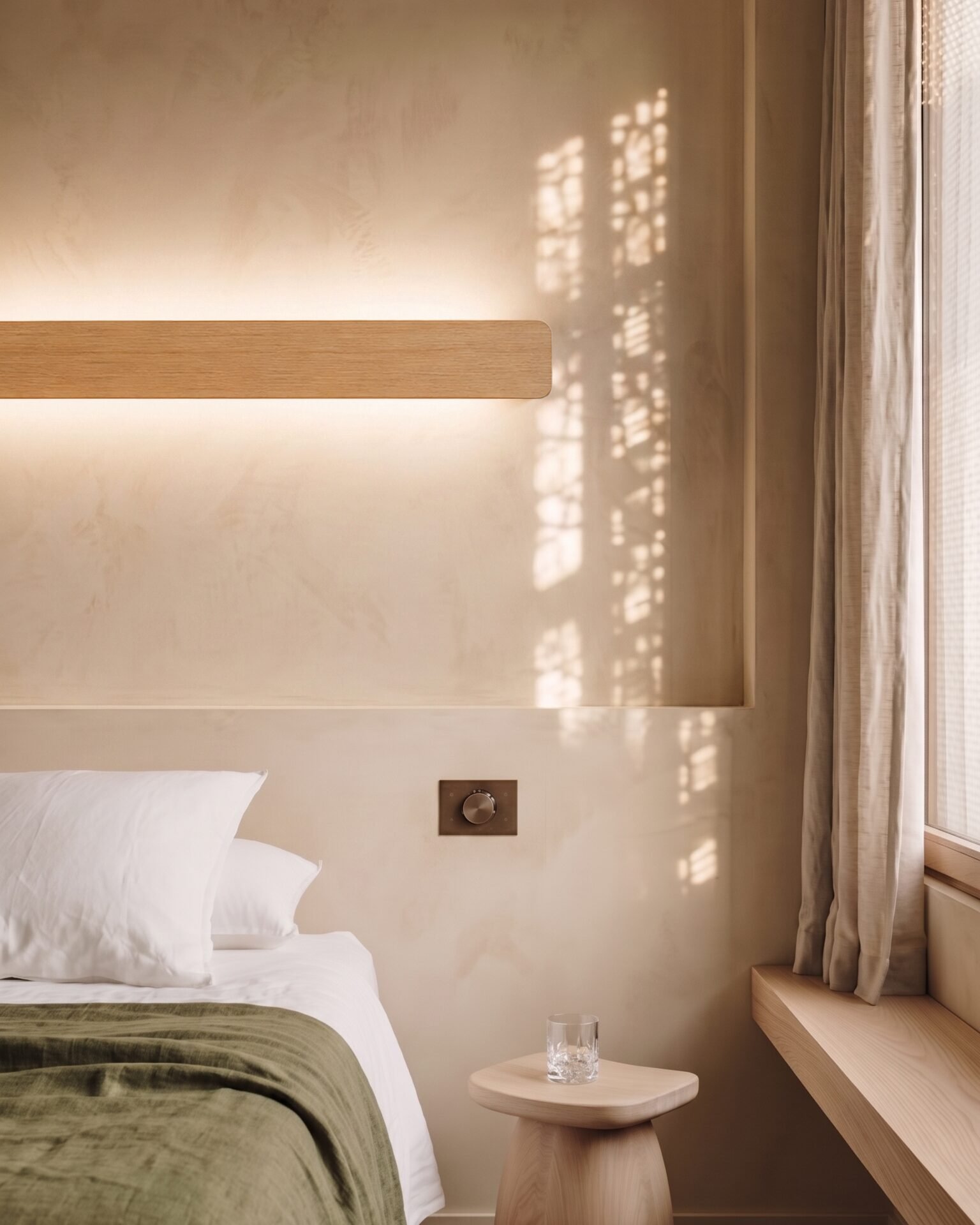 Bedside Nina Wall linear oak up-and-down light by Singular Lighting, next to a dimmer switch on a textured wall. Minimalist bedroom nightstand with a glass of water.