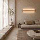 Living area with a low wooden sofa and a Nina Wall linear up-and-down lamp in natural oak, near sheer curtains and a tall vase. Calm, warm ambiance.