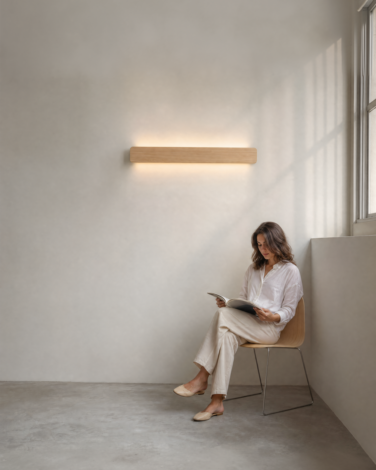 A single 120cm long natural oak Nina Wall W120 linear up-and-down light sconce by Singular Lighting mounted horizontally on a textured wall, casting warm, continuous light over a person reading in a minimalist space.